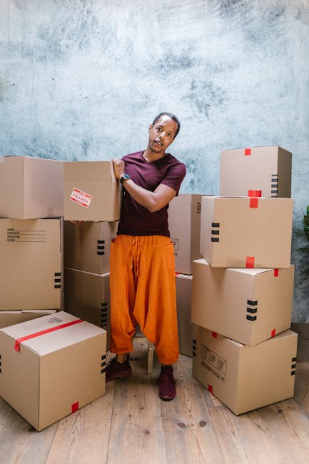 A person with short hair, wearing a dark purple T-shirt, orange trousers, and dark shoes, stands indoors alongside multiple cardboard boxes of varying sizes. Some of the boxes are stacked on the floor