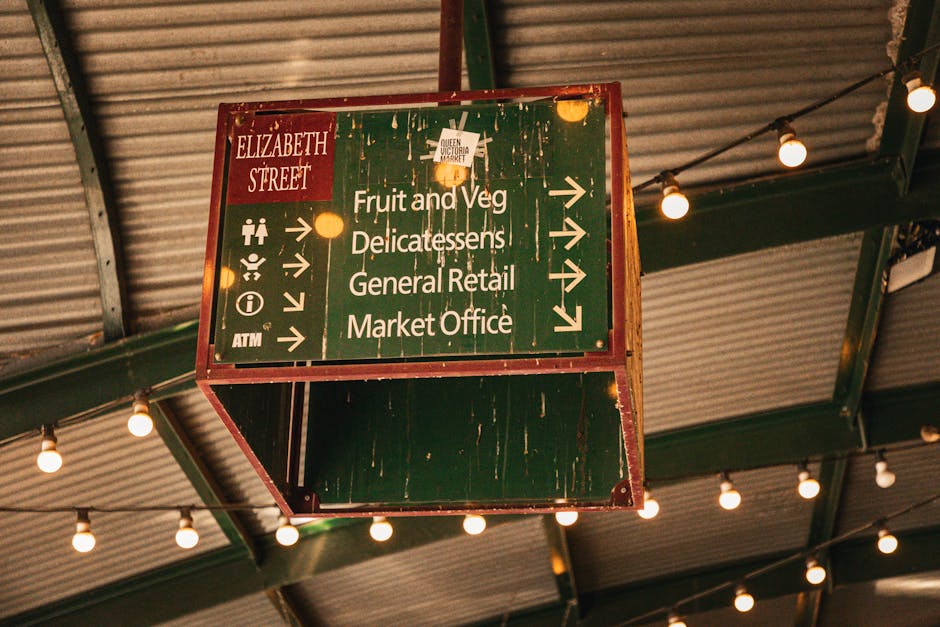 A vintage green directional sign hanging from a metal ceiling structure, displaying white text and icons indicating different sections such as Fruit and Veg, Delicatessen, General Retail, Market Office, and an ATM, all with arrows pointing to the right or left. The sign shows visible signs of wear, with peeling paint and some dirt, and is illuminated by warm string lights attached to the ceiling, creating a well-lit indoor environment likely within a market or shopping area. The ceiling features corrugated metal panels supported by green-painted metal beams, contributing to a rustic, industrial aesthetic. As part of a house removal or relocation process, furniture and equipment, such as loading trolleys or boxes, are not present in this image, but the setting suggests preparation for moving or relocation tasks within a marketplace context, with Brixton Movers possibly involved in the logistical planning.