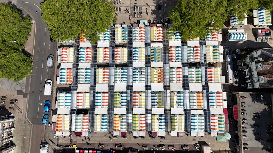 An aerial view of a busy market square with numerous white tents featuring colorful striped canopies in red, blue, yellow, green, and orange, set in a grid-like arrangement. The tents are tightly packed, forming rows and columns, with some tents open, revealing various items inside. Surrounding the market are paved pathways, parked cars, and nearby buildings, including residential and commercial structures. Large, leafy trees border parts of the square, providing shade, and pedestrians are visible walking between the tents and along the adjacent sidewalks. The scene captures the organized layout of a market setup, indicative of packing and moving activities related to house removals or relocation services, as provided by Brixton Movers. The clear daylight highlights the vibrant colors of the tent canopies and the busy nature of the market area, connecting to the themes of furniture transport and home relocation logistics.