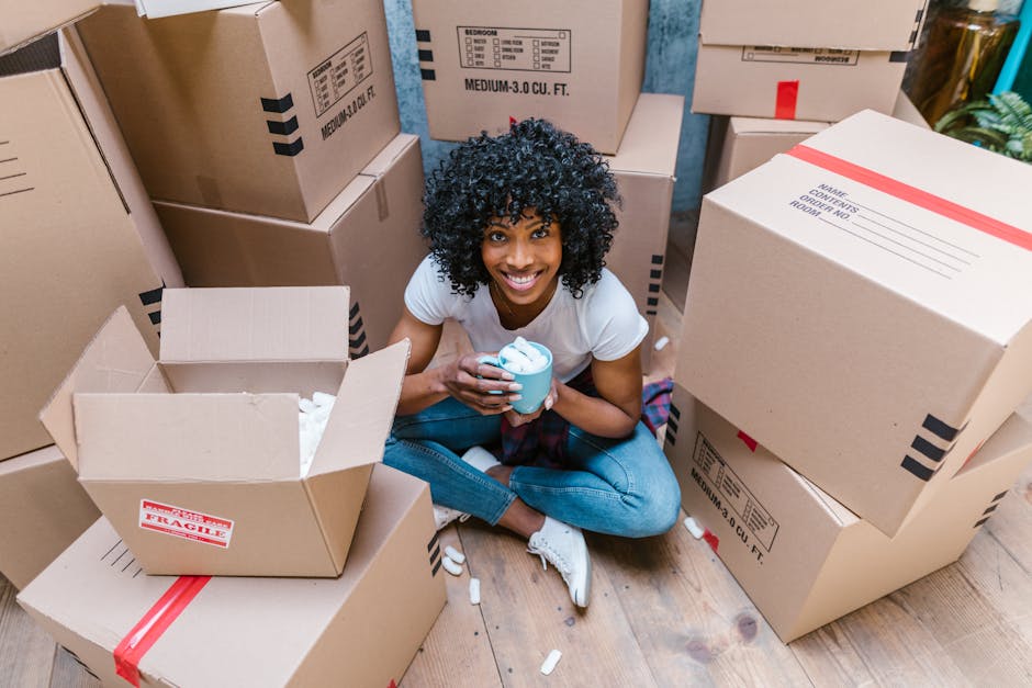 A young woman with curly black hair and a bright smile, sitting on a wooden floor surrounded by various unpacked cardboard boxes, some labeled with fragile or medium-need labels, during a home relocation process. She is holding a disposable coffee cup with foam on top, indicating a break in the packing and moving activity. The scene shows boxes stacked and scattered, with packing materials such as packing peanuts or foam inside some open boxes. The background includes more boxes arranged inside the property, and the lighting appears natural, illuminating the area evenly. This image illustrates the packing stage of a house removal, highlighting careful handling of items, organization with labeled boxes, and a moment of rest amidst a furniture and belongings transport process, reflecting the logistics involved in home relocation services provided by Brixton Movers.