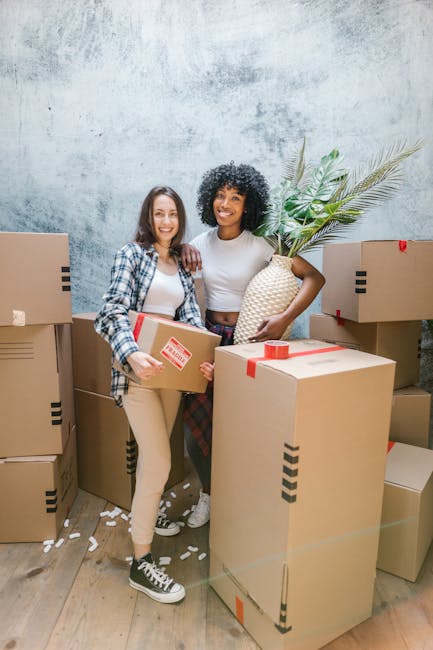 A young woman with curly black hair and a bright smile, sitting on a wooden floor surrounded by various unpacked cardboard boxes, some labeled with fragile or medium-need labels, during a home relocation process. She is holding a disposable coffee cup with foam on top, indicating a break in the packing and moving activity. The scene shows boxes stacked and scattered, with packing materials such as packing peanuts or foam inside some open boxes. The background includes more boxes arranged inside the property, and the lighting appears natural, illuminating the area evenly. This image illustrates the packing stage of a house removal, highlighting careful handling of items, organization with labeled boxes, and a moment of rest amidst a furniture and belongings transport process, reflecting the logistics involved in home relocation services provided by Brixton Movers.